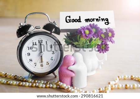 Flowers in a ceramic pots with purple vintage alarm clock and a Good Morning note wooden surface.