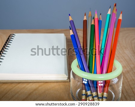 Closeup of color pencils in glass jar with blank notebook on wooden table top