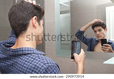 Portrait of attractive young adolescent teenager man using a smartphone device to take selfies pictures of himself in a home bathroom mirror, networking in social media. Technology lifestyle at home.