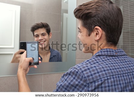 Portrait of a smiling young adolescent teenager man using a smartphone device to take selfies pictures of himself in a home bathroom mirror, networking in social media. Technology lifestyle at home.