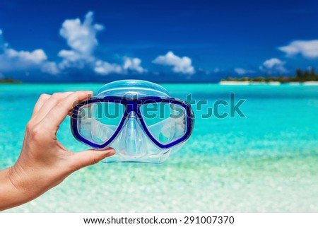 Hand holding snorkel googles against blurred beach and blue sky