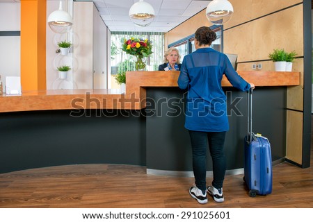 Young woman with a carry on bag at a check in desk, where a female attendant offers information and assistance Royalty-Free Stock Photo #291025601