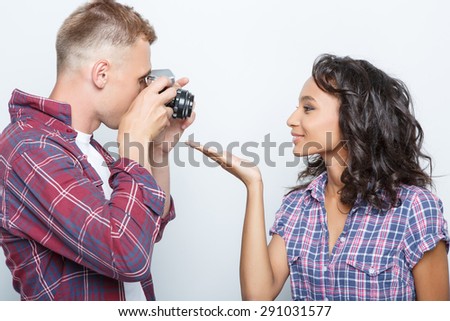 Mixed couple. Beautiful young multiracial girl posing and sending a kiss to her blond boyfriend making a photo on a camera, isolated on grey background