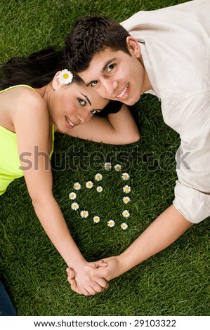 Young couple in love lying in a meadow while looking at camera, heart shape of daisy in the grass
