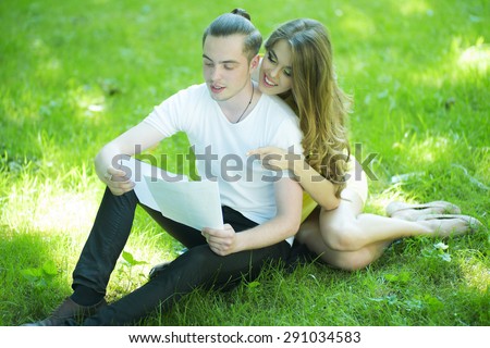 Young handsome man relaxing in park sitting with alluring smiling girl on green grass reading on natural background, horizontal picture