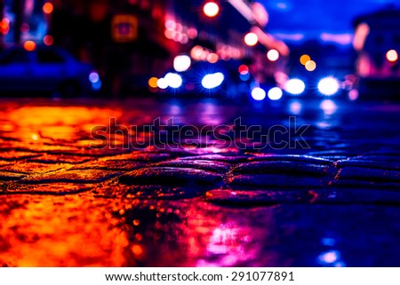 The bright lights of the evening city after rain, headlights from cars in the distance. View from the pavement level, image in the orange-blue toning