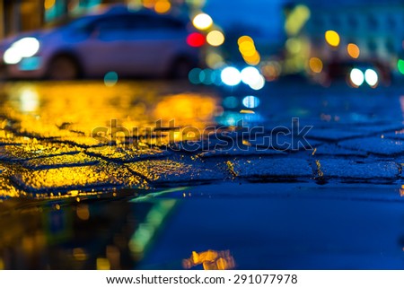 The bright lights of the evening city after rain, headlights of the cars riding straight. View from the pavement level next to the roadside puddle