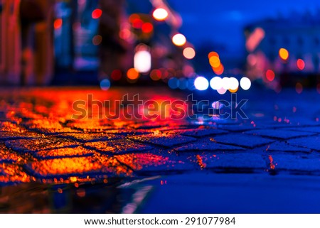 The bright lights of the evening city after rain, headlights of the cars riding straight. View from the pavement level next to the roadside puddle, image in the red-blue toning
