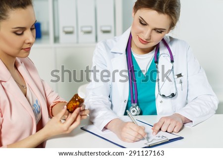 Beautiful young female medicine doctor sitting in front of working table and writing prescription on special form while patient holding jar of pills. Medical and pharmacy concept