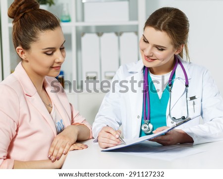 Beautiful smiling female medicine doctor holding clipped pad with document and pointing in it with pen to patient. Signing treatment bill or insurance policy concept Royalty-Free Stock Photo #291127232