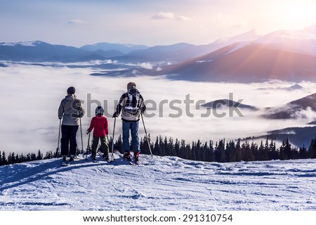 People observing mountain scenery.
Family of three people stays in front of scenic landscape. These are skiers, they dressed in winter sport jackets and have skies attached.
