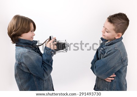 Twins photo shoot.Young child instant camera photographer takes a picture of and young child boy model posing on a white background. Both are wearing denim jackets.