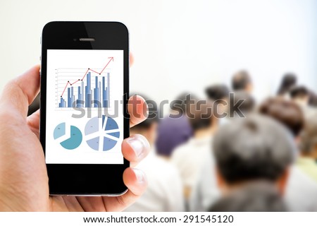 Close-up hand holding mobile phone with analyzing graph against people in business conference and presentation background 