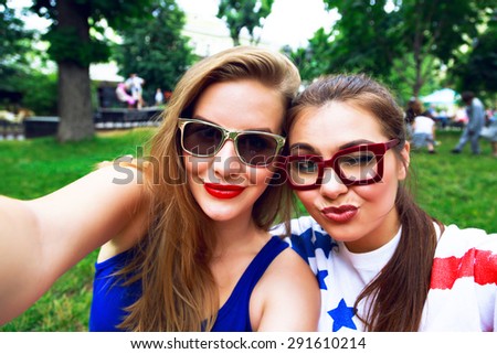 Outdoor lifestyle portrait of two sisters making selfie, smiling and winking, bright clothes and make up. Best friends having fun at city park, joy, happiness.