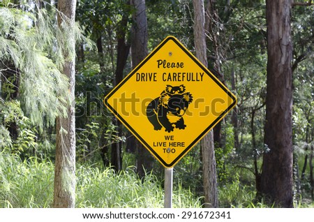 Warning sign for motorists about koala habitat. Bright yellow diamond shape sign with mother and baby koala. Writing on sign: Please drive carefully we live here too. Trees behind sign. 