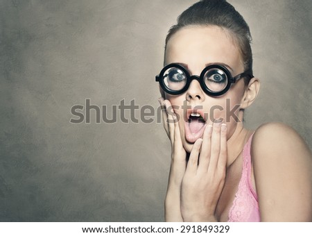 Shock grimace of young pretty woman in funny round glasses on grey background
