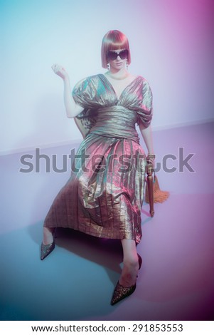 Full Length View of Glamorous Woman with Red Hair Wearing Sunglasses and Shiny Retro Gown Sitting in Chair Looking Bored and Holding Fringed Purse in Smoky Disco Night Club Setting