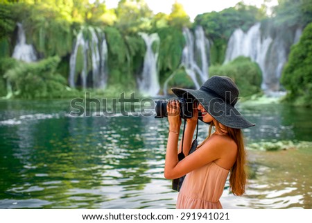 Young woman tourist in pink dress and big hat photographing with professional photo camera beautiful Kravica waterfall in Bosnia and Herzegovina