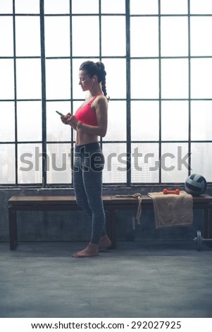 Standing by a wooden bench which is placed by a big window, a fit, healthy, strong young woman is looking down at her device and selecting which music to listen to for her workout.