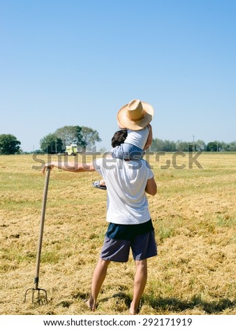 Picture of father on field holding pitchfork riding his son on shoulders wearing big hat looking at combined harvester over blue sky background copyspace