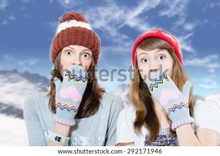 Two surprised young girls in hats and mittens covering mouth with hand  on mountains background
