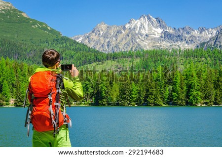 Young man with an orange backpack takes a photo of a mountain lake, Strbske Pleso, High Tatras, Slovakia