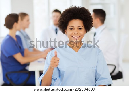 clinic, profession, people and medicine concept - happy female doctor or nurse over group of medics meeting at hospital showing thumbs up gesture