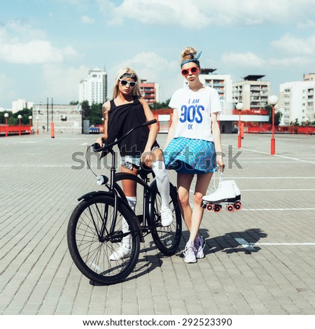 Outdoor fashion portrait of attractive young women with bicycle and roller skates. Two teen swag girls