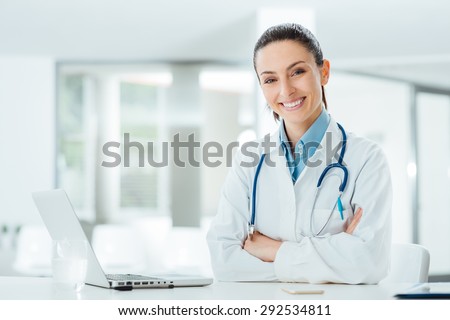 Confident female doctor sitting at office desk and smiling at camera, health care and prevention concept Royalty-Free Stock Photo #292534811