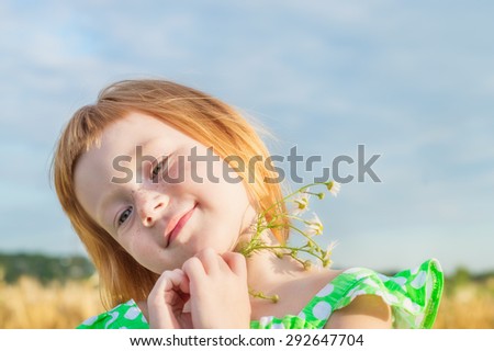 funny red-haired girl holding daisies in hands on a background of a wheat field and blue sky