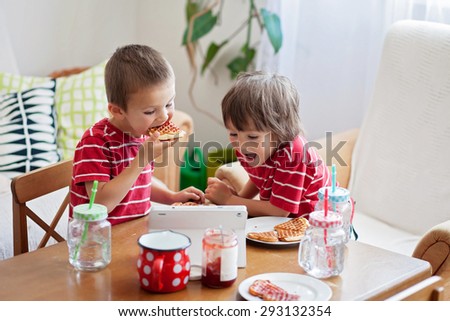 Two happy kids, two brothers, having healthy breakfast sitting at wooden table in sunny kitchen, eating waffles  and watching cartoon on tablet