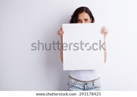 Attractive young girl is showing a white square. It closes half of her face. She looking forward confidently. Isolated on white and copy space on right