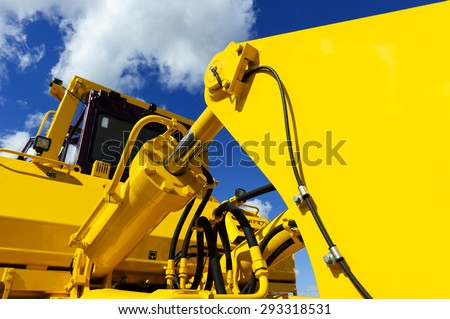 Bulldozer, huge yellow powerful construction machinery with big bucket, focused on hydraulic piston arm, blue sky and white clouds on background Royalty-Free Stock Photo #293318531