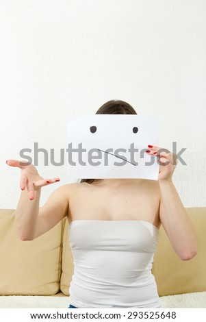 Young woman is confused. Girl doesn't know what to do. Girl holds forefinger and smiley drown on paper. Young  female bears sad face drawing on white sheet. Wonering about things.