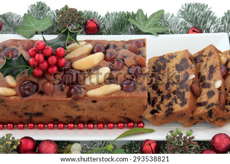 Genoa cake and slices with holly, red bauble christmas decorations and winter greenery over white background.
