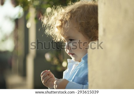 Small cute smiling baby boy with blond curly hair standing in street near wall on outdoor background, horizontal picture