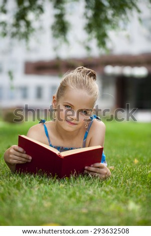 Portrait of beautiful happy little girl reading red book lying on green grass in the summer park