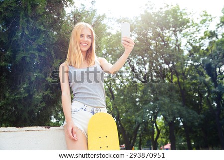 Smiling young girl with skateboard making selfie photo on smartphone outdoors