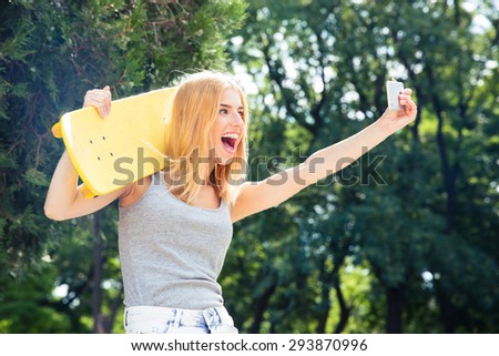 Funny young girl with skateboard making selfie photo on smartphone outdoors