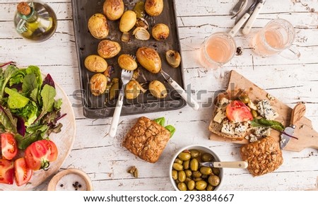 Summer lunch served on a wooden picnic table with fruits, various cheese, vegetables, leaves of salad, olives, fruits juice  and oven sheet with cooked potatoes from above. Rustic mediterranean food.