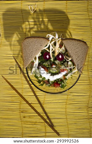 Tomatoes and cucumbers in a glass plate with chopsticks, a piece of rye bread on a yellow bamboo mat on a wooden background, studio lights lit, the view from the top.
