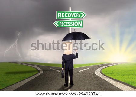 Worker with cardboard head and umbrella, standing on the road while looking at signpost directing at the road to recovery or recession financial