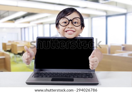 Portrait of pretty little girl smiling on the camera with empty laptop screen and showing thumbs up in the class