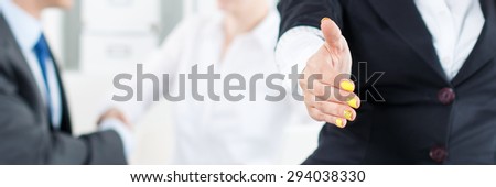 Beautiful smiling business woman in suit offering hand to shake while couple employees working in background. Serious business and partnership concept. Formal greeting and welcoming gesture