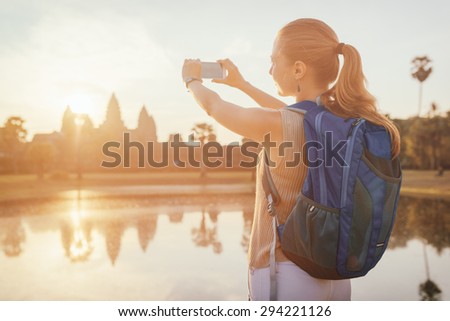 Young female tourist taking picture with smartphone of the ancient temple complex Angkor Wat reflected in a pond at sunrise on background of rising sun. Siem Reap, Cambodia. Toned image.