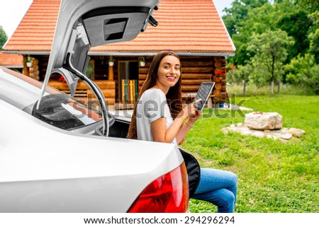 Young and attractive woman packing with digital tablet near the car with wooden house on background.
