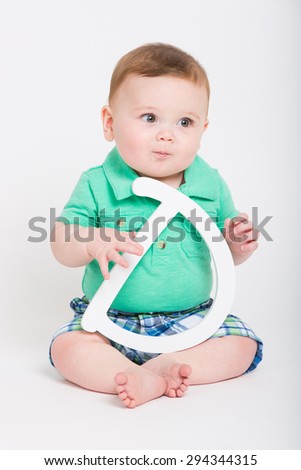 8 month year old baby sits on a white background holding a white letter D looking off camera. dressed in a cute green polo shirt and blue plaid shorts.