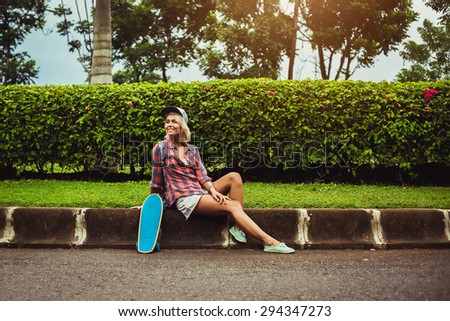 Skateboarder girl sit on border in cap and checkered shirt with her skateboard