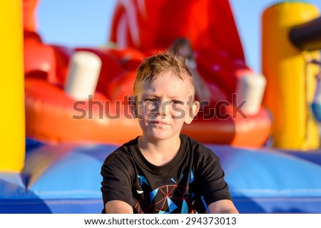 Thoughtful serious little boy in a kids playground sitting in the evening sunshine in front of a colorful jumping castle