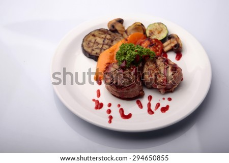 Delicious pieces of juicy grilled tenderloin wrapped bacon drizzled sauce with chopped grilled paprika eggplant zucchini tomato onion mushrooms parsley on plate isolated on white, horizontal picture